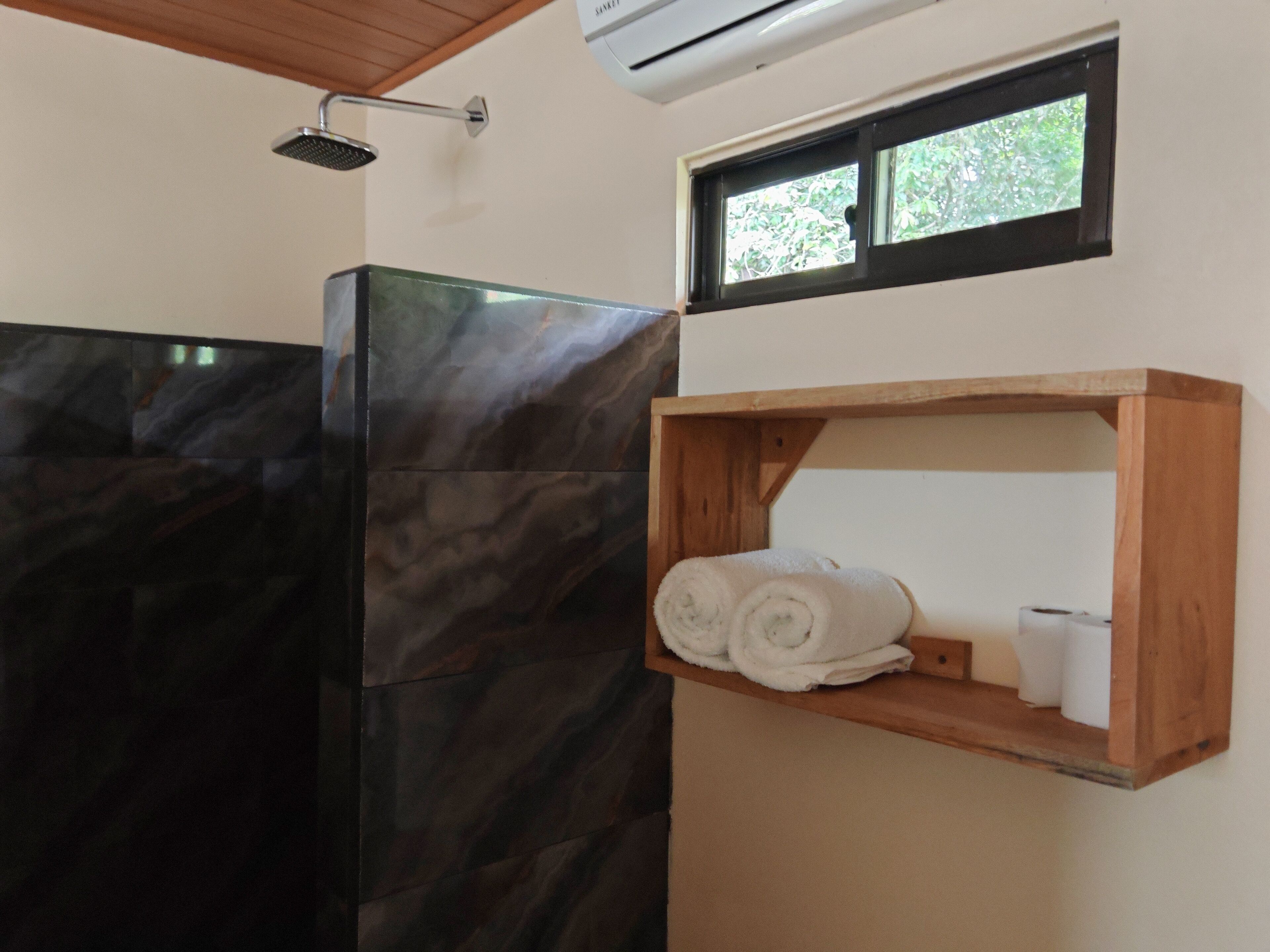 A bathroom with a black marble shower enclosure, a wooden shelf with towels and toilet paper, and a window with a view of trees.