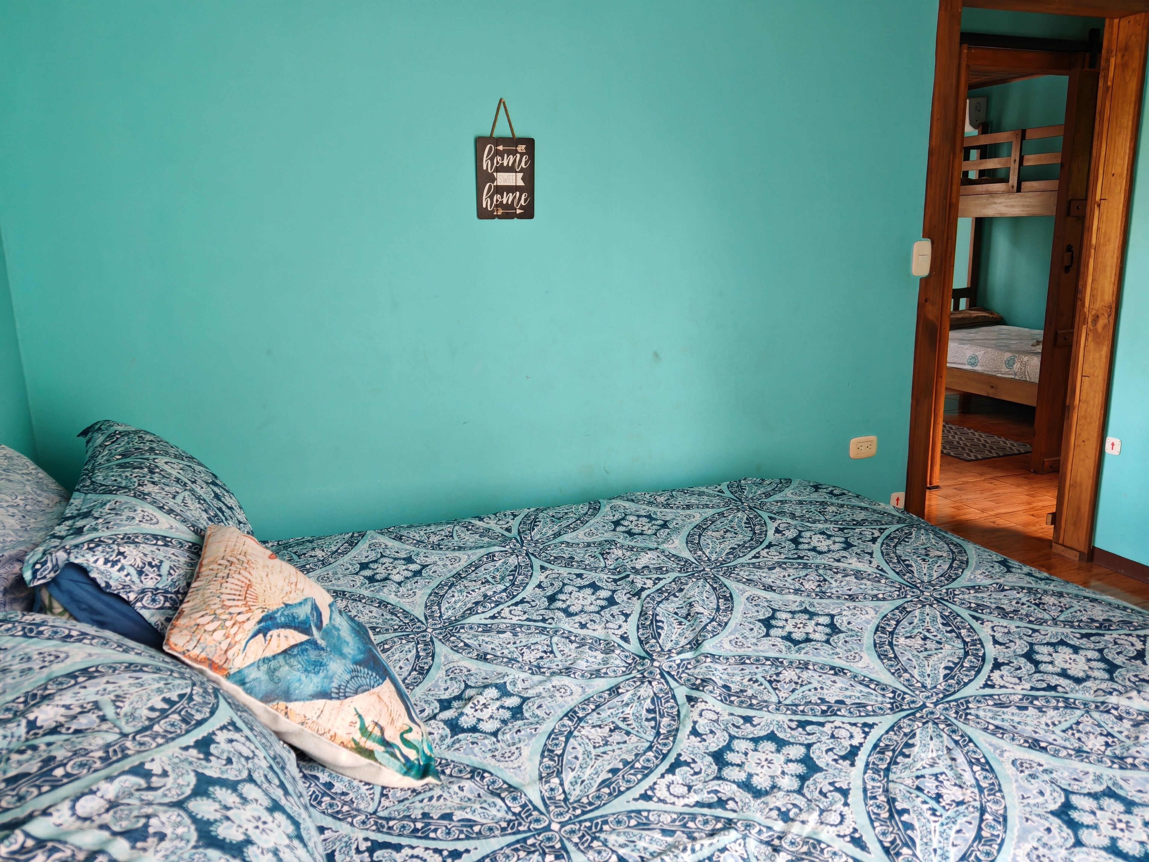 A room with a bed featuring a blue and white patterned bedspread, a hanging sign on the wall, and a wooden door leading to another room.