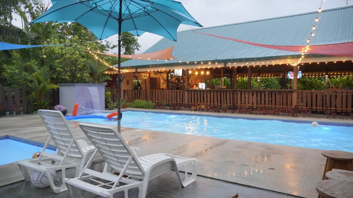 Un área de piscina con sillas de descanso, un espacio de estar cubierto y luces colgantes.