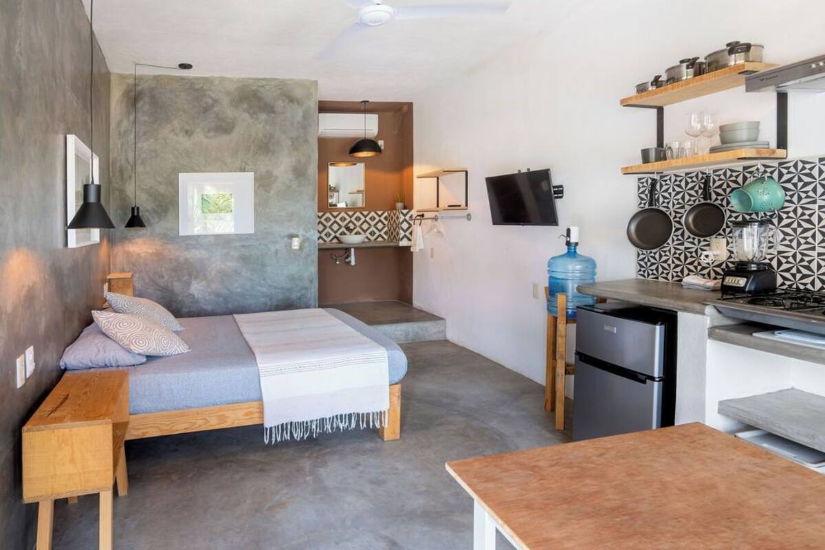 A compact, modern kitchen with a built-in stove, a sink, and a countertop. There is a bed with a wooden frame and a gray mattress in the adjacent room.