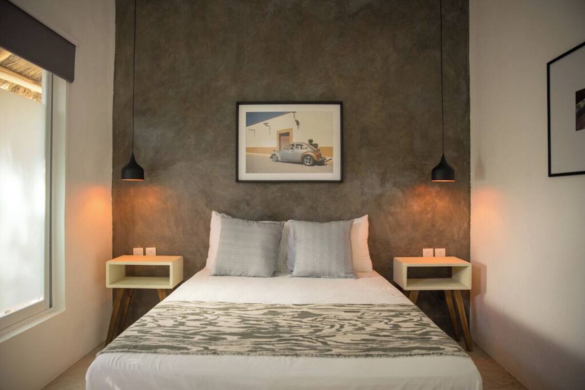 A bedroom with a large bed, two bedside tables, a framed picture on the wall, and a window on the left.