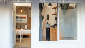 Quarto com cama, escrivaninha e cadeira, cozinha equipada com fogão, e uma janela grande com vista para o exterior.
