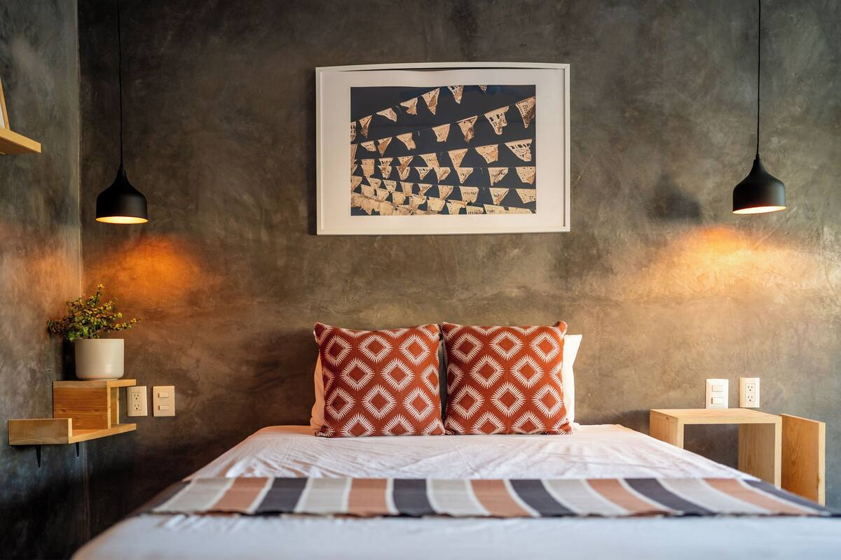 A modern bedroom with a bed, two wall-mounted pendant lights, a framed artwork, and a small wooden nightstand with a plant.