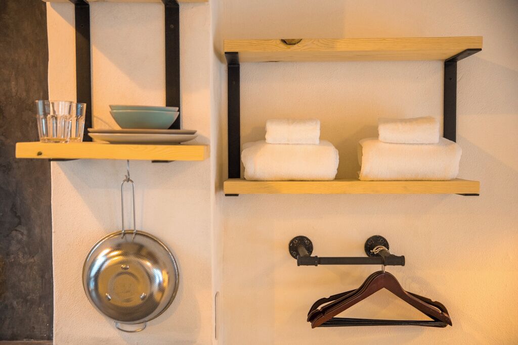 Una repisa montada en la pared con un colador metálico, una repisa de madera con toallas dobladas y un perchero de madera.