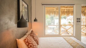 Quarto com cama, quadro emoldurado, luminária pendente e janela com vista para área externa com telhado de palha.