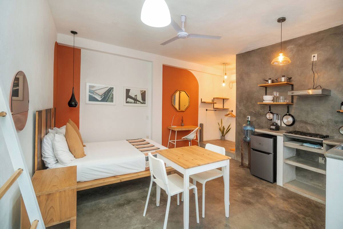 A compact, modern room with a bed, a wooden table, and a kitchenette.
