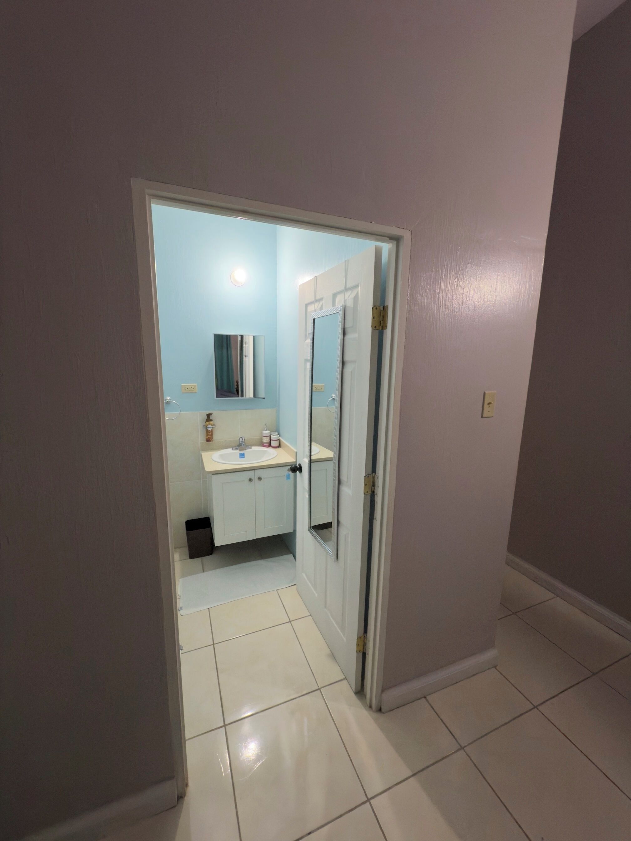 Een badkamer met witte tegels, een wastafel, een spiegel en een deur naar een andere kamer.
