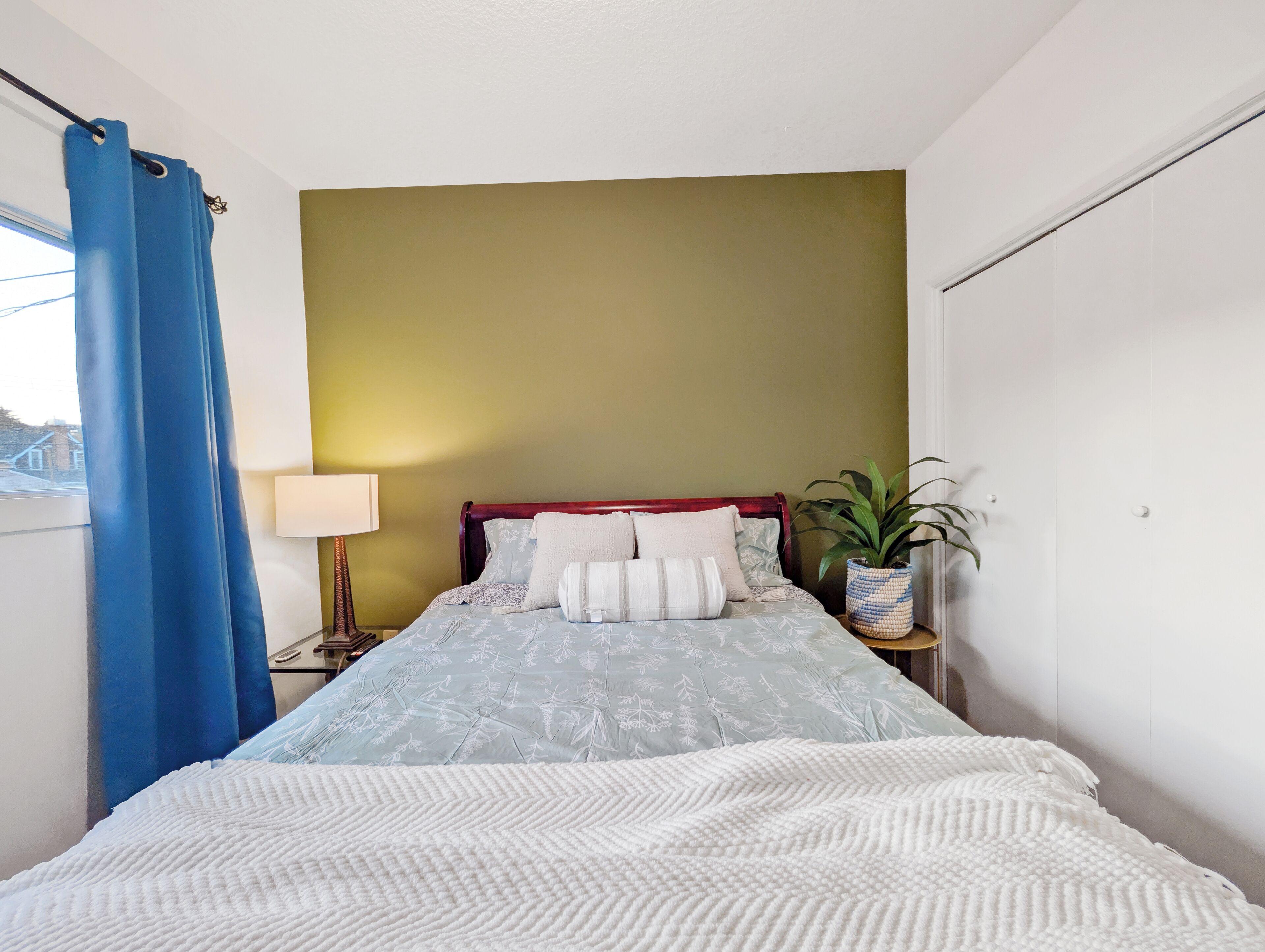 Une chambre à coucher avec un lit, une table de chevet, une lampe, une plante en pot et une fenêtre avec des rideaux bleus.