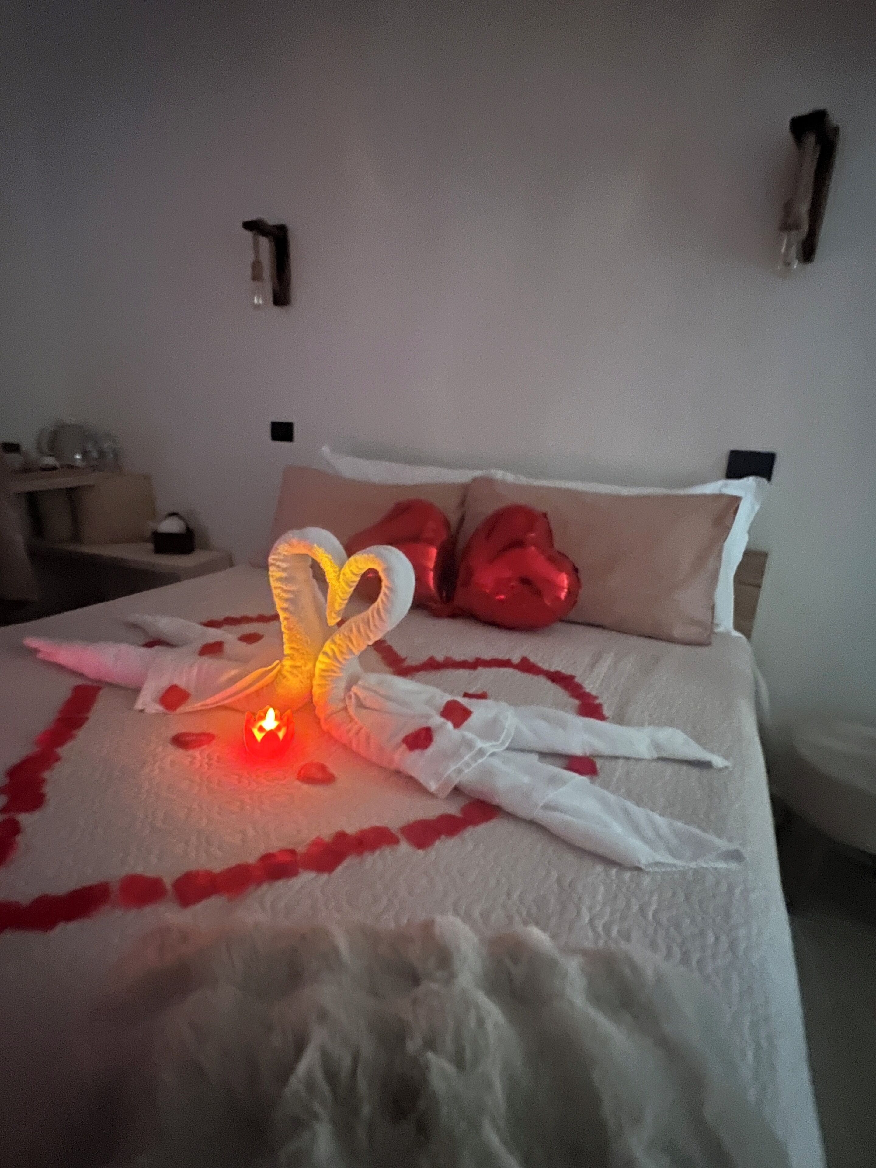 Ein Bett mit einer herzförmigen Anordnung aus roten Rosenblüten, einem weißen Stoff und einer brennenden Kerze.