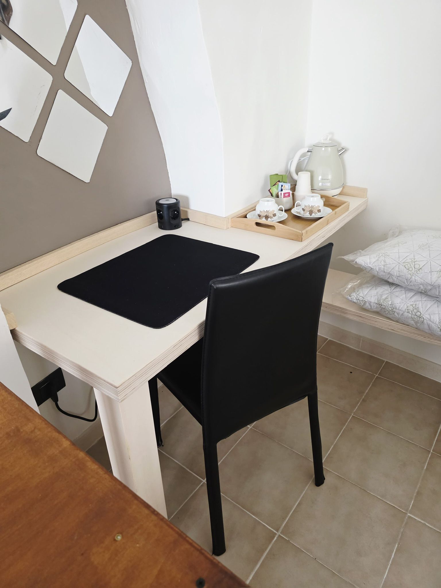 Un espacio de trabajo pequeño y ordenado con escritorio, silla y estante con tetera, tazas y bandeja.