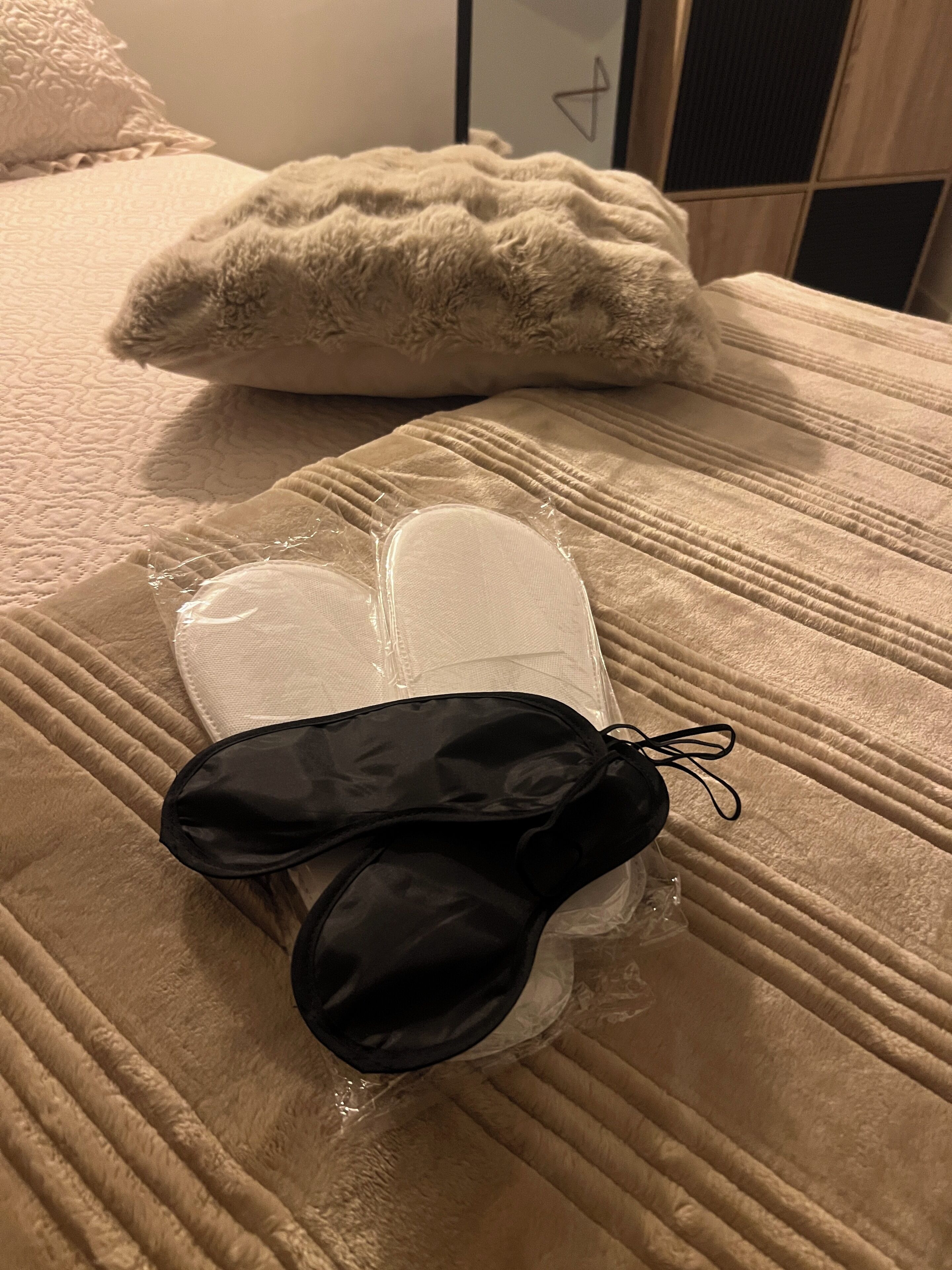 Una cama con cubrecolcha color beige, una almohada blanca y un objeto negro envuelto en plástico transparente.