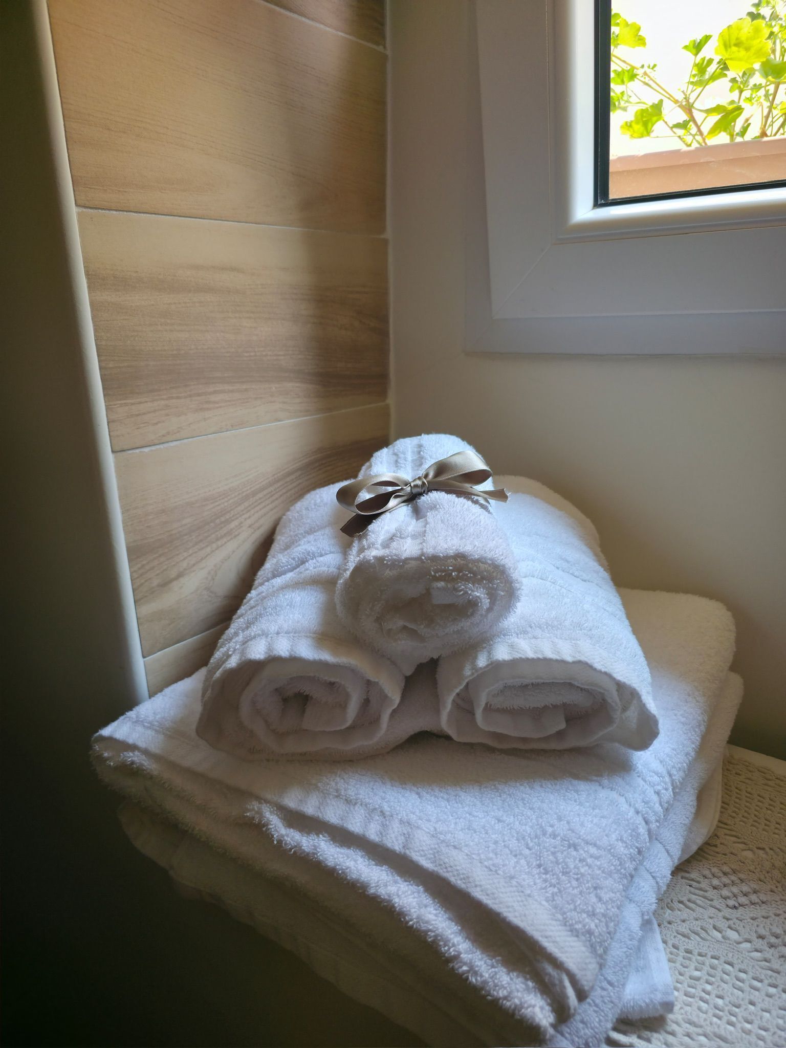 A neatly folded white towel with a ribbon on a white surface, possibly a bathroom counter, with a wooden wall and a window with a view of greenery.