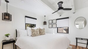 A bedroom with a large bed, a ceiling fan, a hanging light fixture, a bedside table, a mirror, and a window with a view. - The Coral House — Modern Luxury 1BR with Ocean View Balcony | Cal King Bed + Queen Sleeper, Sleeps 4 · Folding Glass Balcony Doors · Full Kitchen · AC · Two Exclusive Beach Cruisers · Avila Beach · Managed by Solmaré Stays (Avila Beach)