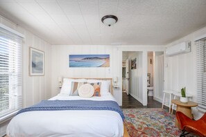 A bedroom with a bed, a nightstand, a window with blinds, a painting on the wall, and a hallway leading to a bathroom. - The Pine House — Dog Friendly Studio with Ocean View Deck | Queen Bed Under the Pines · In-Unit Washer & Dryer · AC · Full Kitchenette · Beach Gear & Bikes · Avila Beach · Sleeps 2 · Managed by Solmaré Stays (Avila Beach)