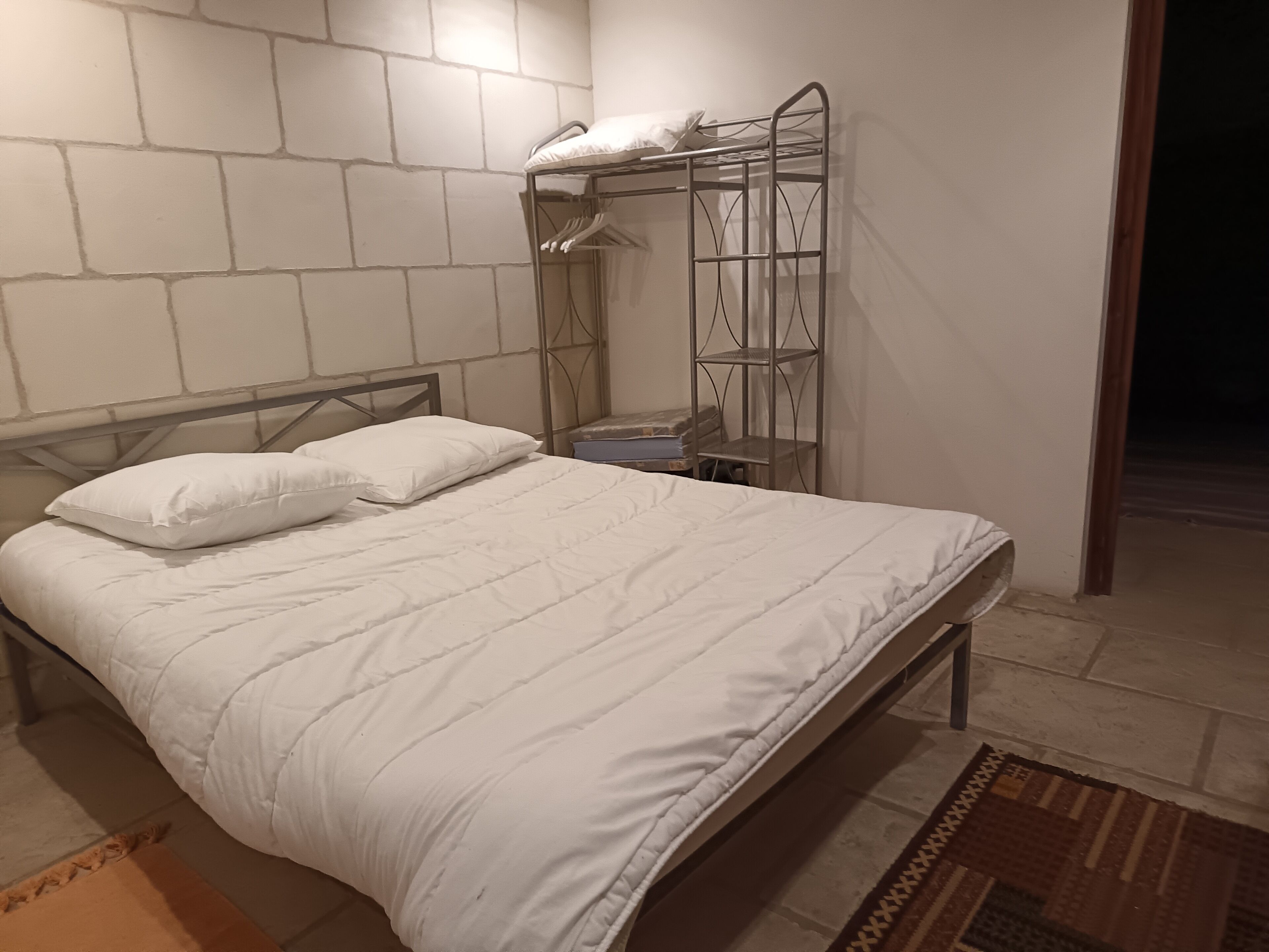 A neatly made bed with white bedding in a room with a brick wall and a metal shelf unit.