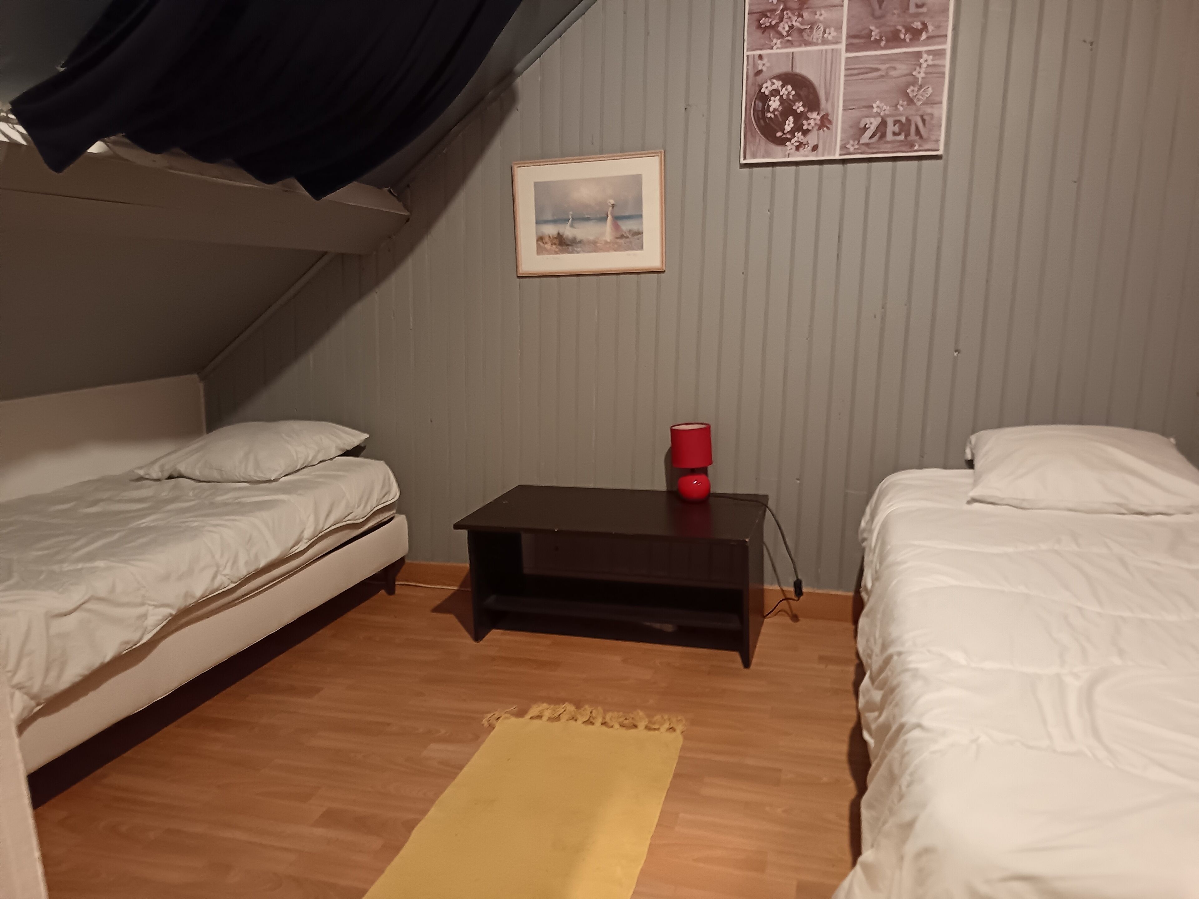 A small, cozy room with two beds, a wooden floor, a small table with a red lamp, and a framed picture on the wall.