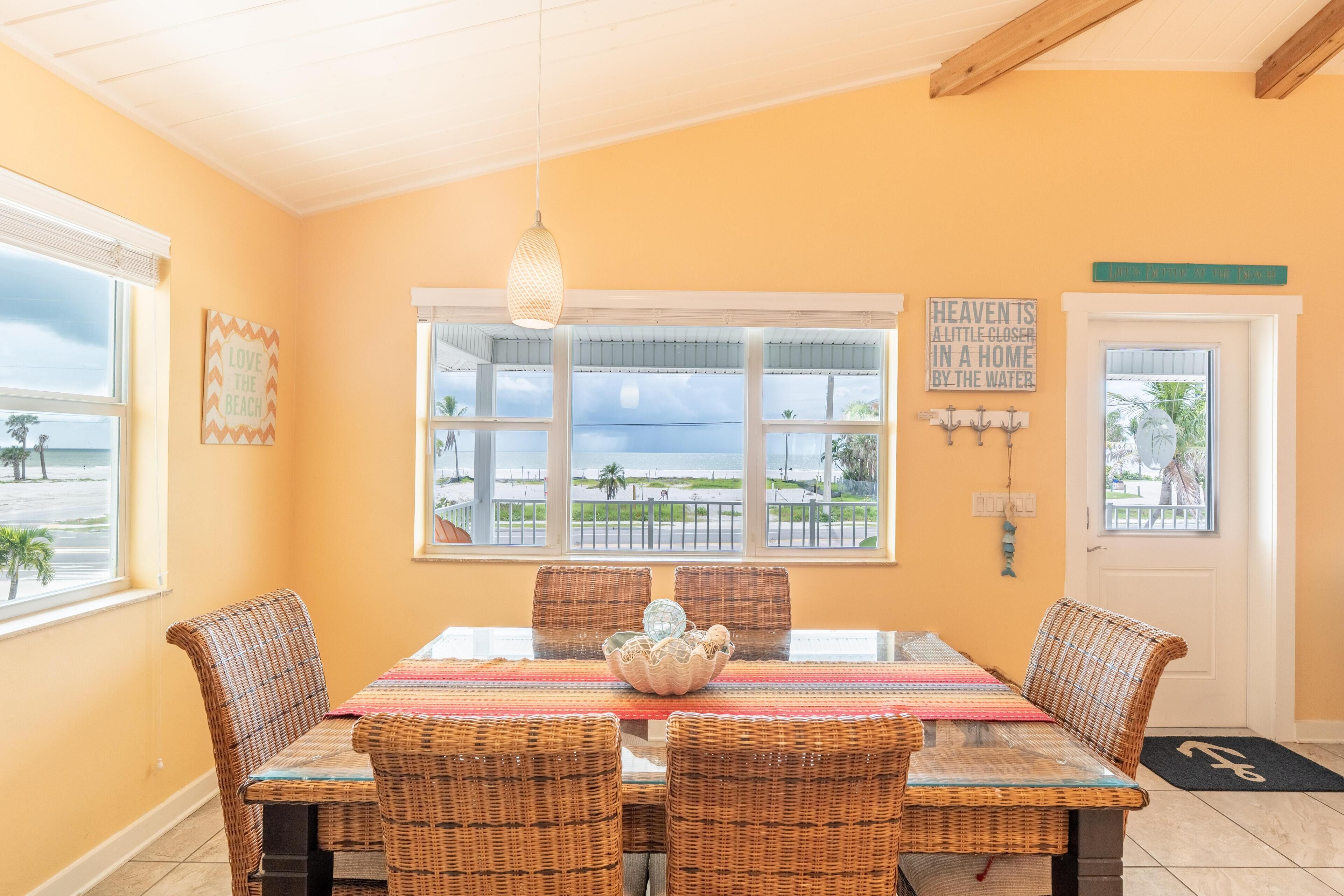 A dining area with wicker chairs, a glass table, and a view of the beach through large windows.