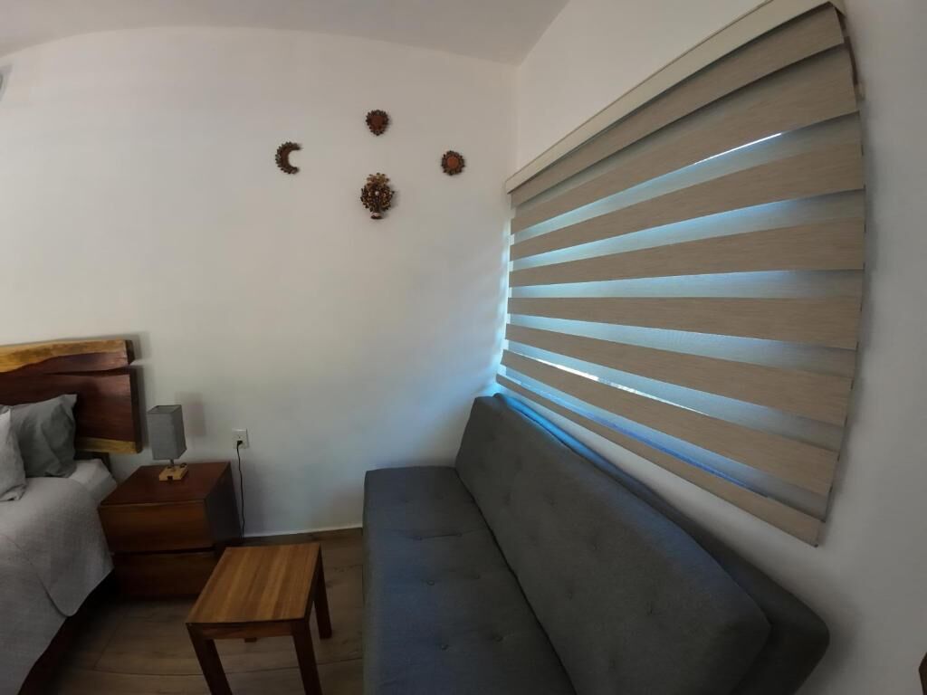 A modern living room with a grey sofa, a wooden coffee table, and a wooden nightstand with a lamp. There are wall-mounted decorations and a window with horizontal blinds.