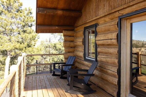 Una cabaña de troncos con un balcón de madera, dos mecedoras y una ventana con vistas a un bosque.