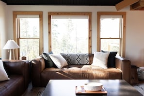 A room with a brown leather sofa, a wooden table, and three large windows with wooden frames.