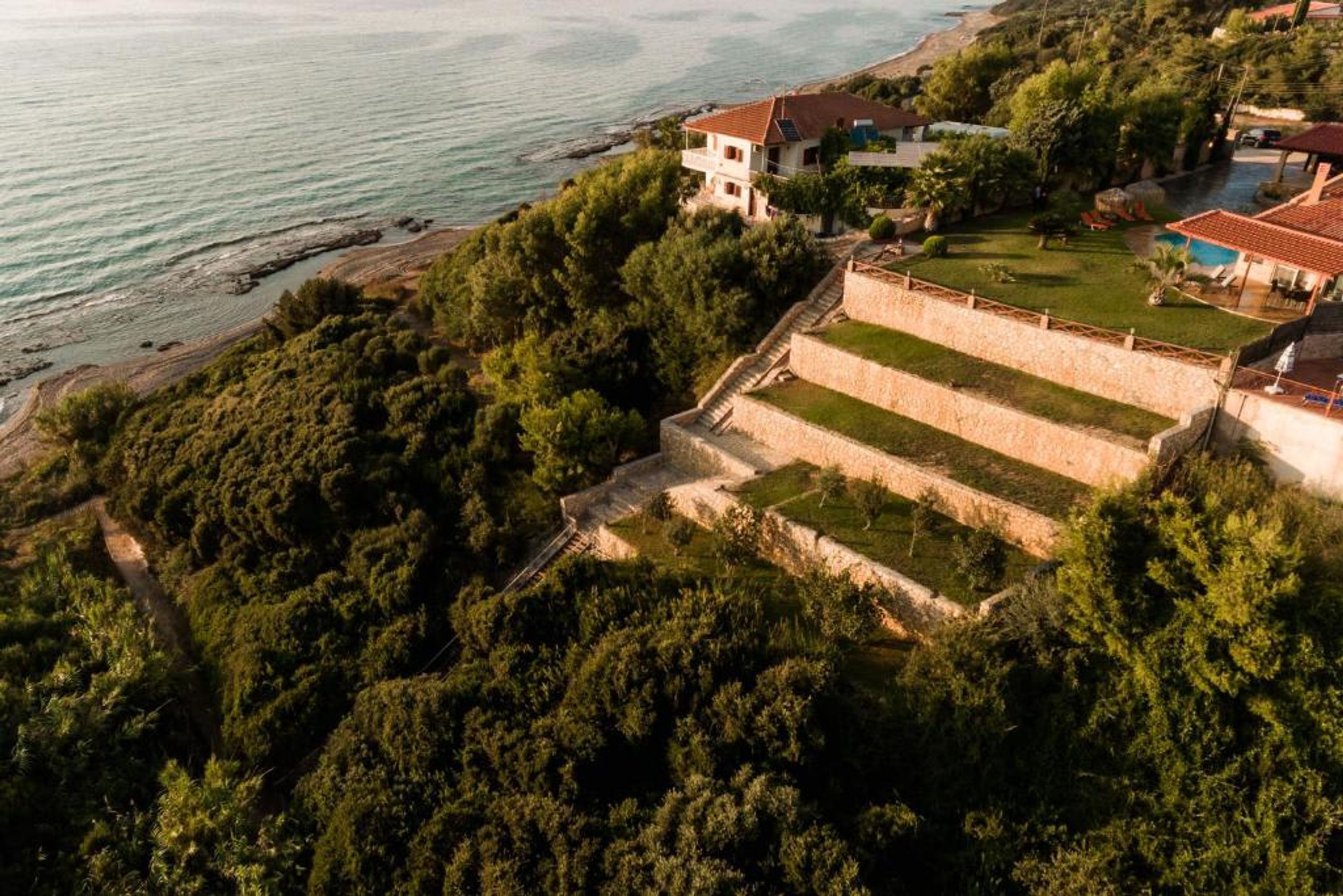 An aerial view of a coastal property with a terraced garden, a beach, and a building with a red roof.