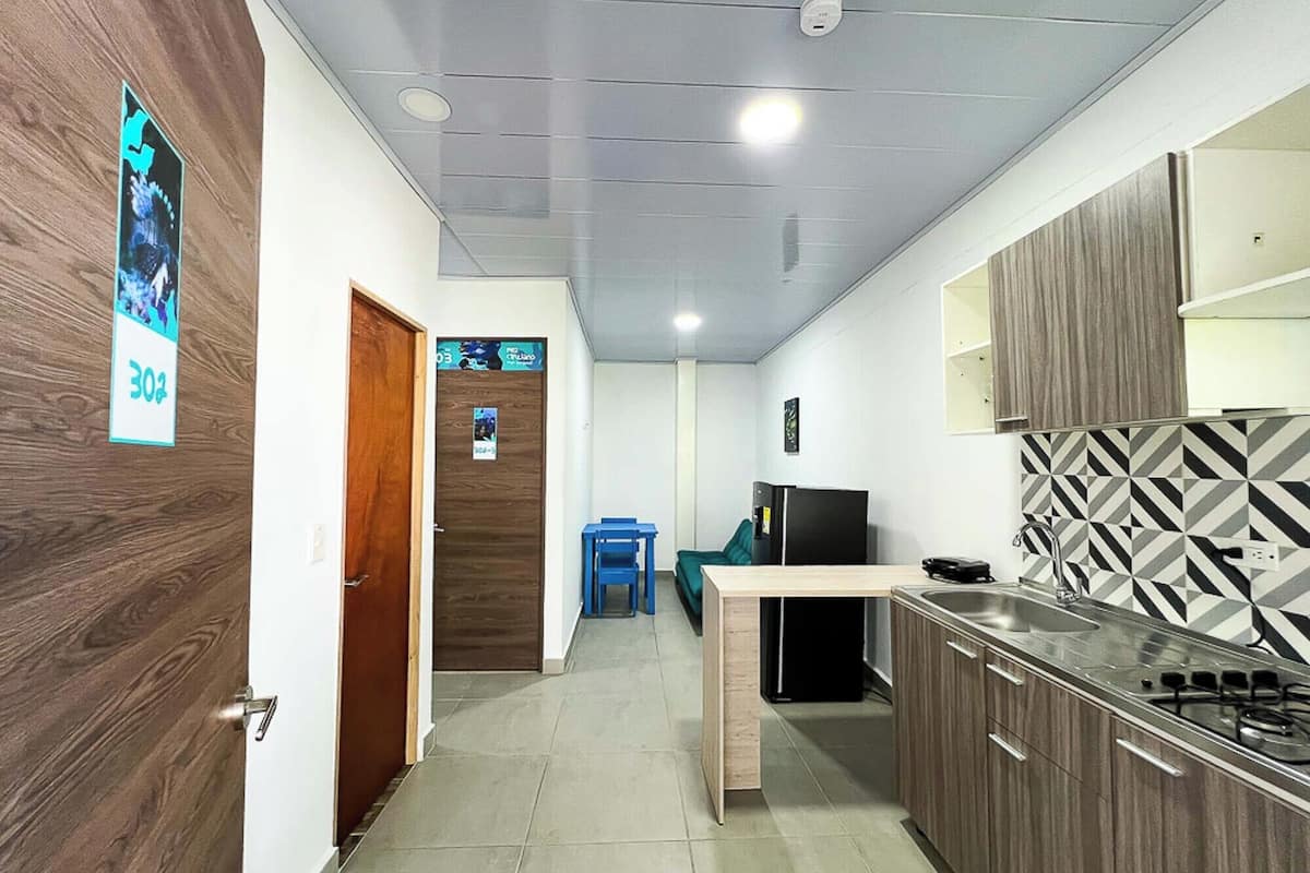 A compact kitchen with a built-in stove, a refrigerator, and a microwave. There is a wooden door on the left and a blue chair in the background.