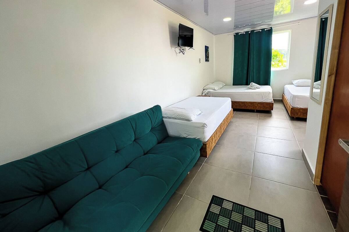 A room with a teal sofa, two single beds, a TV mounted on the wall, and a window with curtains.