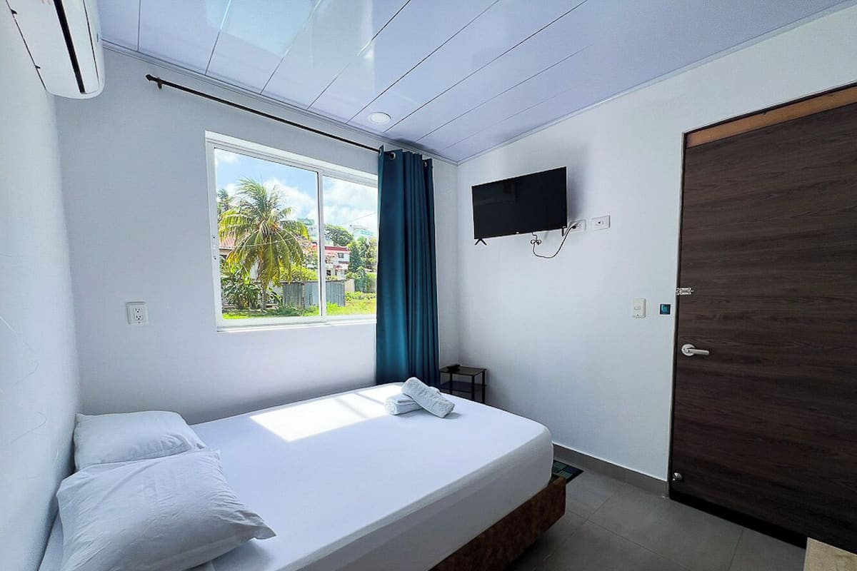 A bedroom with a bed, a window with curtains, a TV mounted on the wall, and a door.
