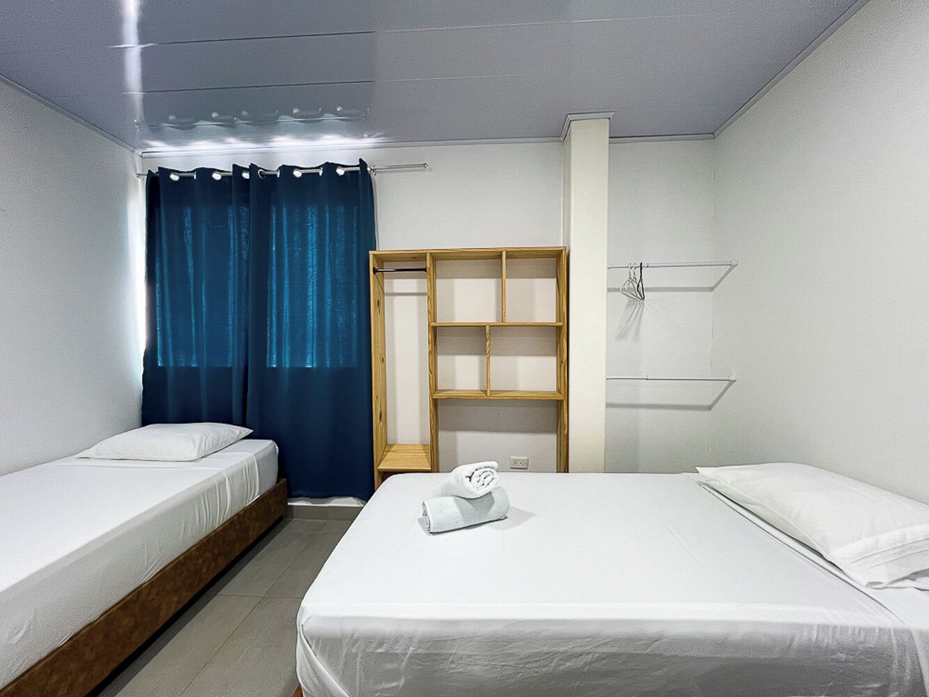 A hotel room with two single beds, a wooden wardrobe, a small table, and a blue curtain.