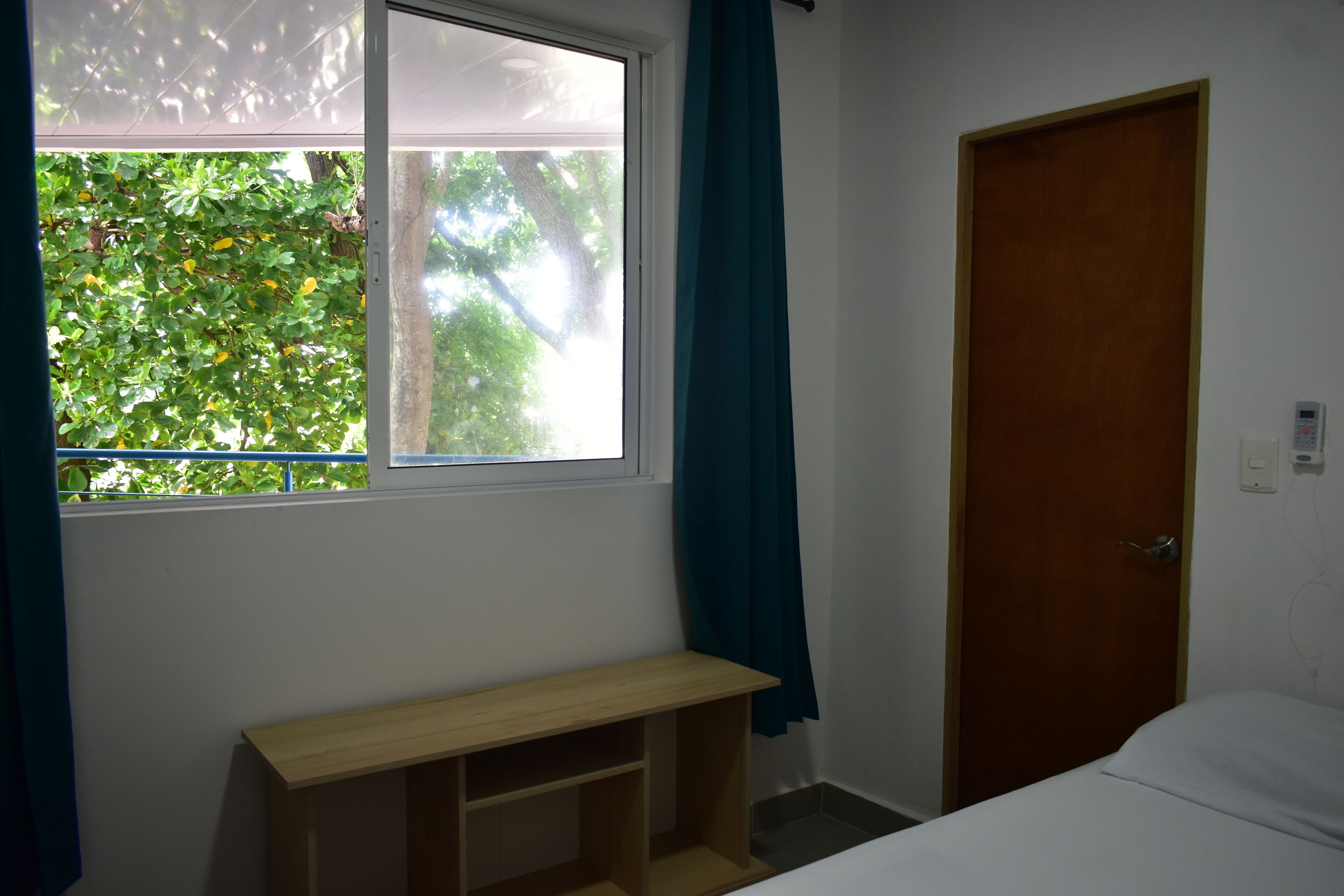 A room with a bed, a window with curtains, a wooden shelf, and a door.