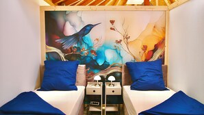 Une chambre à coucher avec deux lits, chacun recouvert d’une courtepointe bleue, et un grand tableau mural représentant un oiseau et des fleurs.