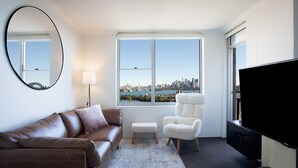 A modern living room with a leather sofa, a white armchair, a small stool, a large TV, and a window with a cityscape view.