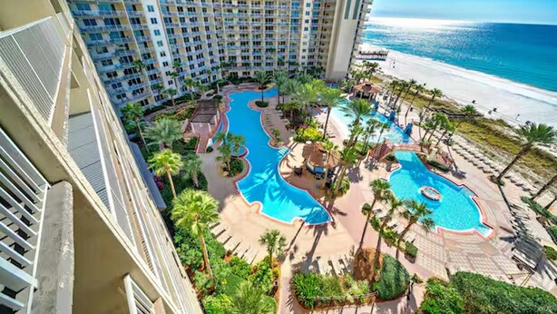 A high-rise hotel with multiple balconies overlooking a beach and a pool area with palm trees and lounge chairs. - Beach, Please! (Panama City Beach)