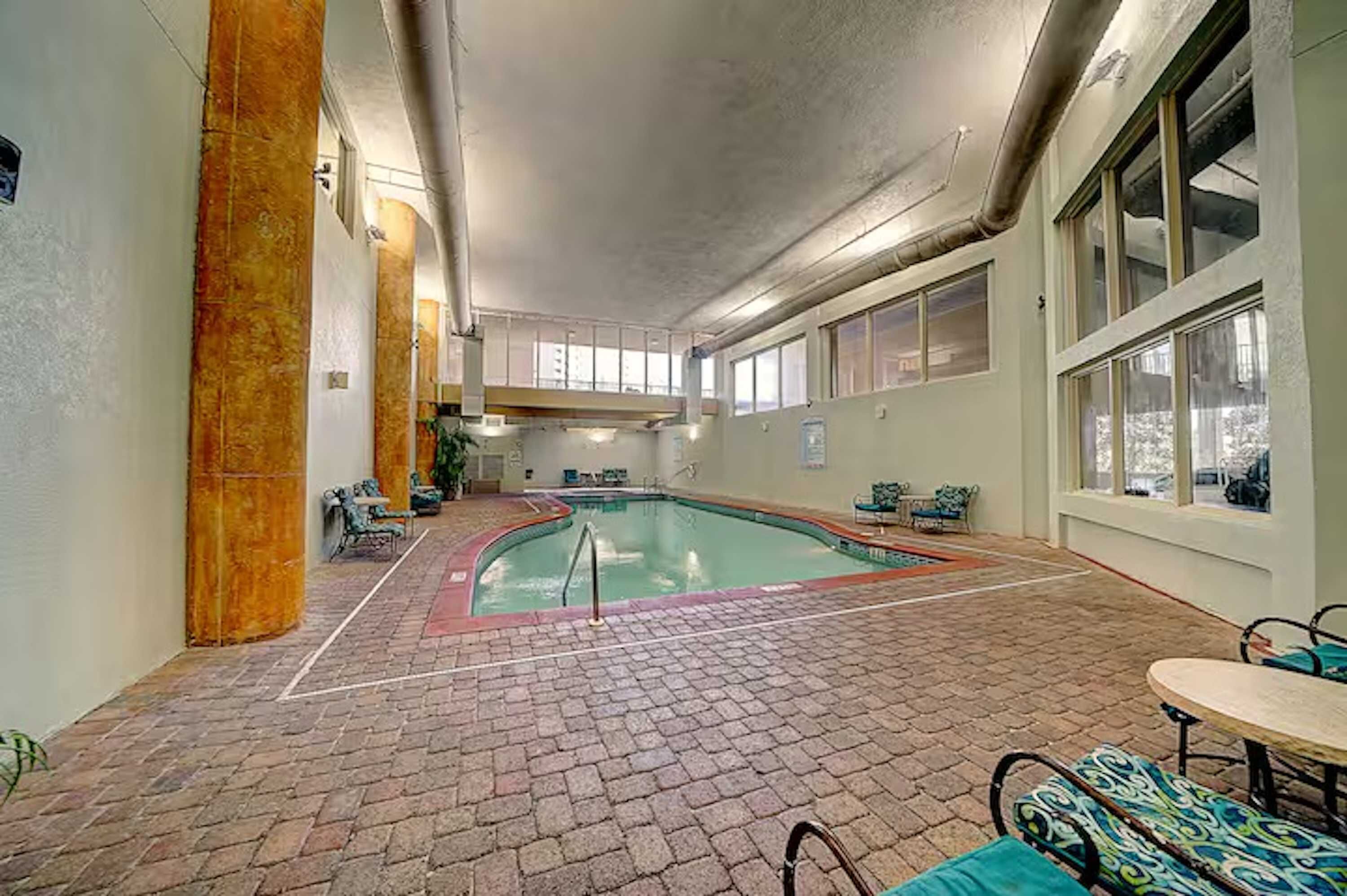 An indoor swimming pool with tiled flooring, seating areas, and large windows.