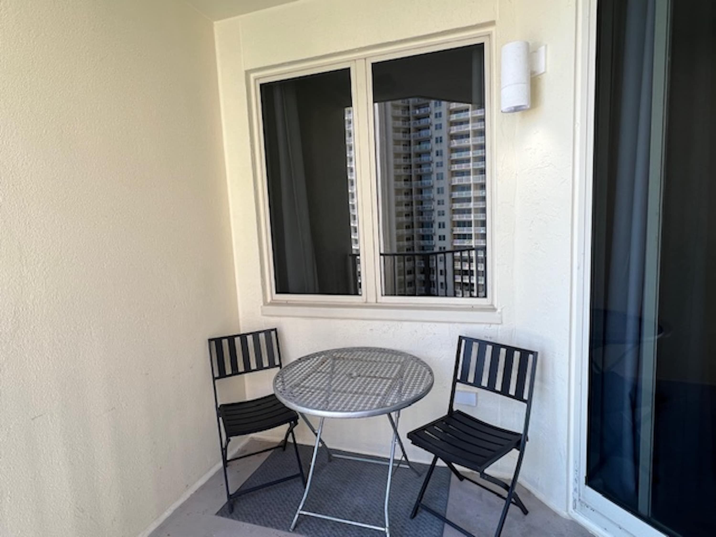 Un piccolo balcone con un tavolino rotondo e due sedie, una finestra con tende, e vista su un grattacielo.