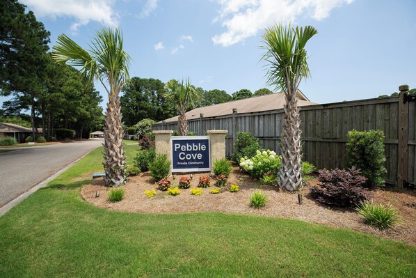 A sign for Pebble Cove Private Community with a wooden fence, palm trees, and landscaped garden.