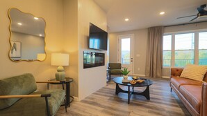 A modern living room with a green sofa, a round coffee table with snacks, a TV on the wall, and a balcony with curtains. - 3BR Condo Near Ripken Experience & LeConte Center (Pigeon Forge)