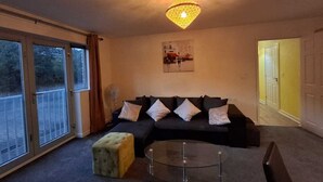 A living room with a black sectional sofa, a glass coffee table, and a yellow ottoman. There is a large window with curtains, a ceiling light, and a door leading to another room.
