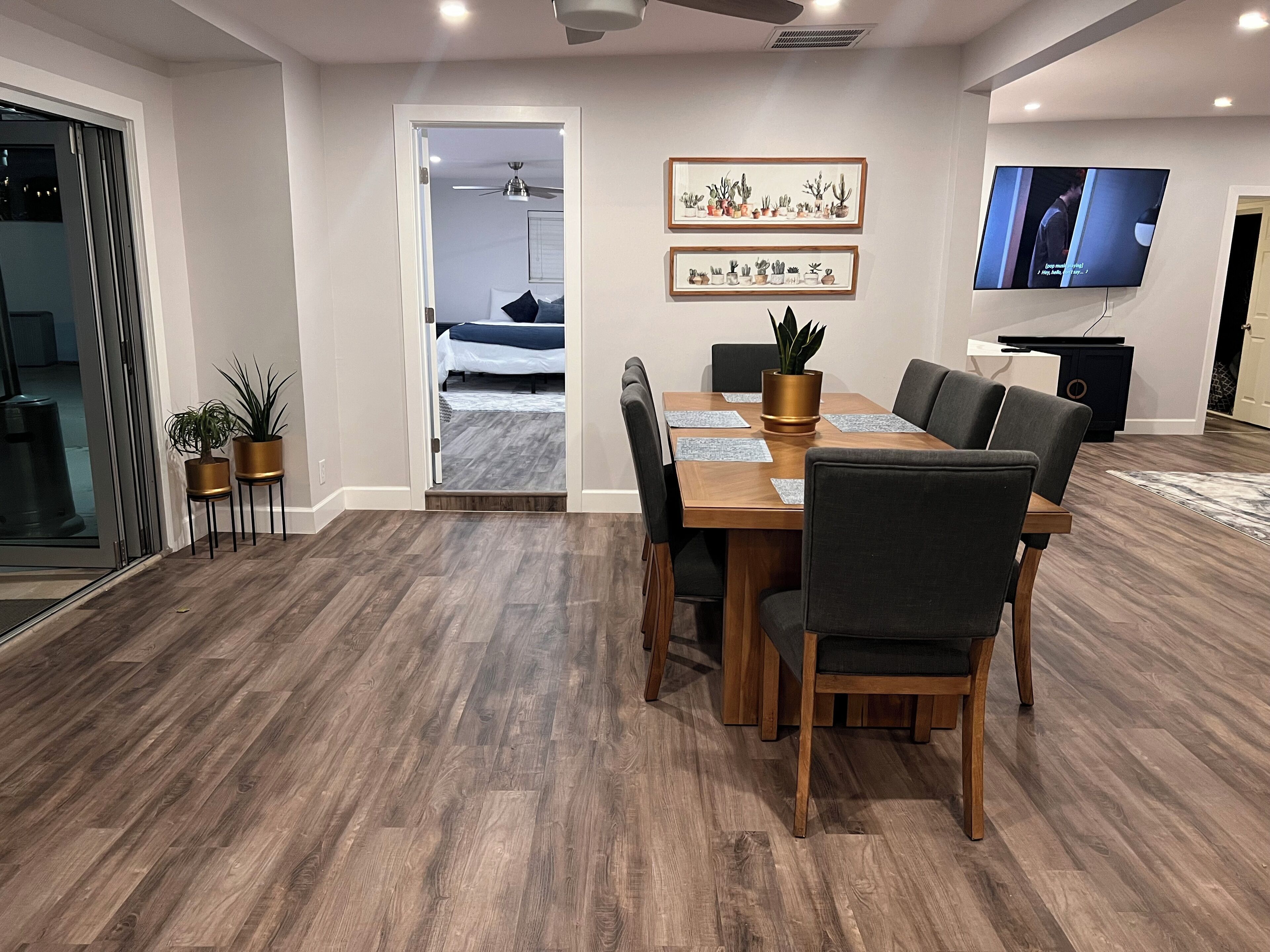 A modern dining area with a wooden table and chairs, a television mounted on the wall, and framed cactus artwork.