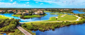 A golf course with water bodies, a wooden bridge, and residential buildings in the background.