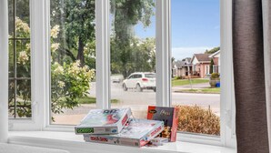 Una ventana con vista a una calle residencial, un coche blanco y un alféizar con juegos de mesa.