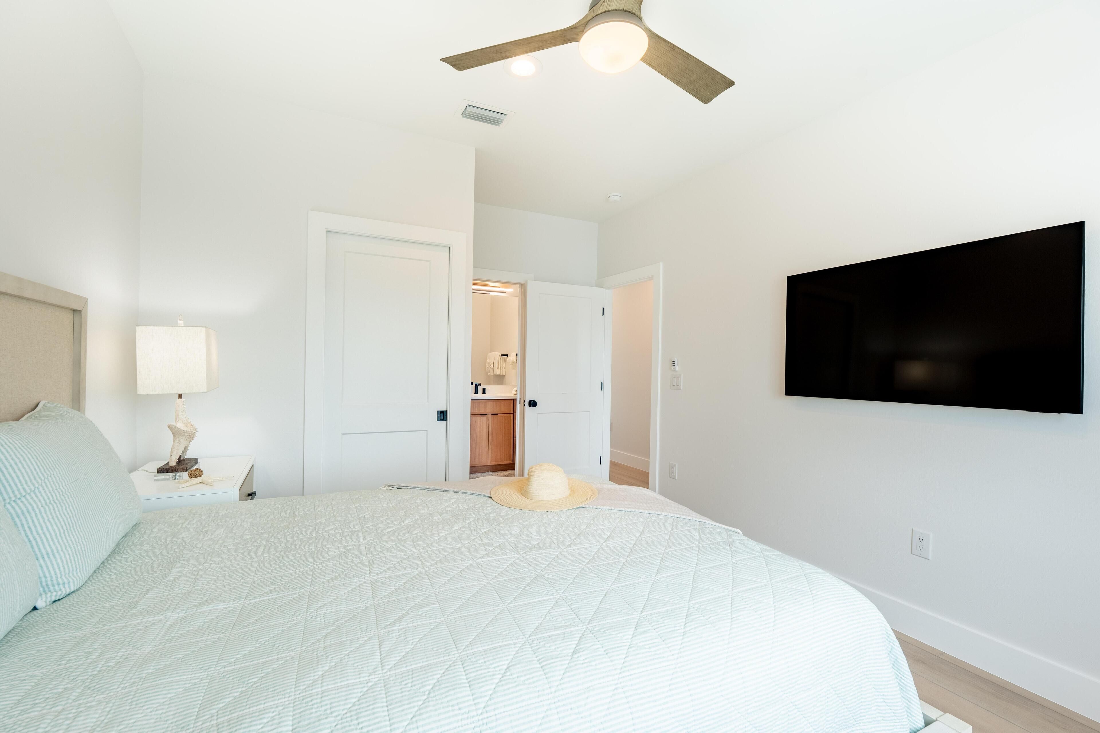 Une chambre à coucher comprenant un lit, un ventilateur de plafond, une télévision et une porte donnant accès à une salle de bain.