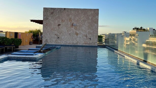 Una piscina infinita en la azotea con un muro de piedra, terraza de madera y cielo azul despejado.