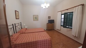 Una habitación con dos camas, un pequeño aparador de madera, un televisor encima, una araña y una ventana con cortinas.