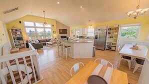 A spacious kitchen with a central island, a dining area with a wooden table, and a living area with a sofa and bookshelves. - 7BR Walk-to-Beach Corolla Home • Hot Tub • Family-Friendly • Sleeps 16+ (Corrolla)