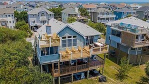 A two-story house with a balcony, surrounded by other houses, and a view of the ocean. - 7BR Walk-to-Beach Corolla Home • Hot Tub • Family-Friendly • Sleeps 16+ (Corrolla)