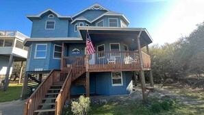 A two-story house with a wooden staircase leading to a balcony, an American flag, and a sign that reads "Sail Away". - 7BR Walk-to-Beach Corolla Home • Hot Tub • Family-Friendly • Sleeps 16+ (Corrolla)