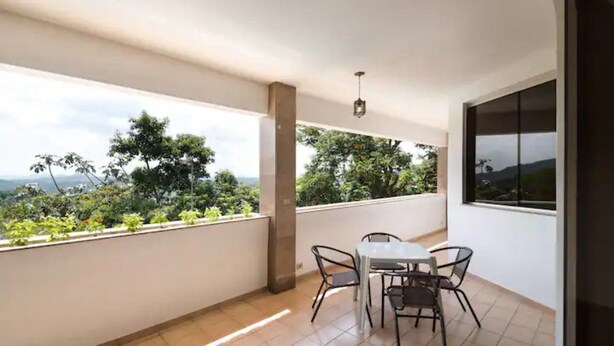 Un balcón con mesa y sillas, una lámpara colgante, y vistas a zonas verdes y colinas.