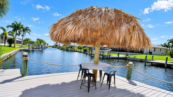 A wooden deck with a thatched umbrella, two chairs, and a table by a calm water body, surrounded by greenery and a clear sky.