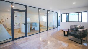 A modern interior with a glass partition labeled "KIDS CLUB," a seating area with a bench and a sofa, and a large window providing natural light. - Ipana by The Spot Rentals (Playa del Carmen)